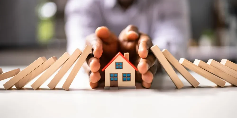 home_insurance Hands protecting a small model house surrounded by falling wooden blocks, symbolizing homeowners insurance coverage and protection.