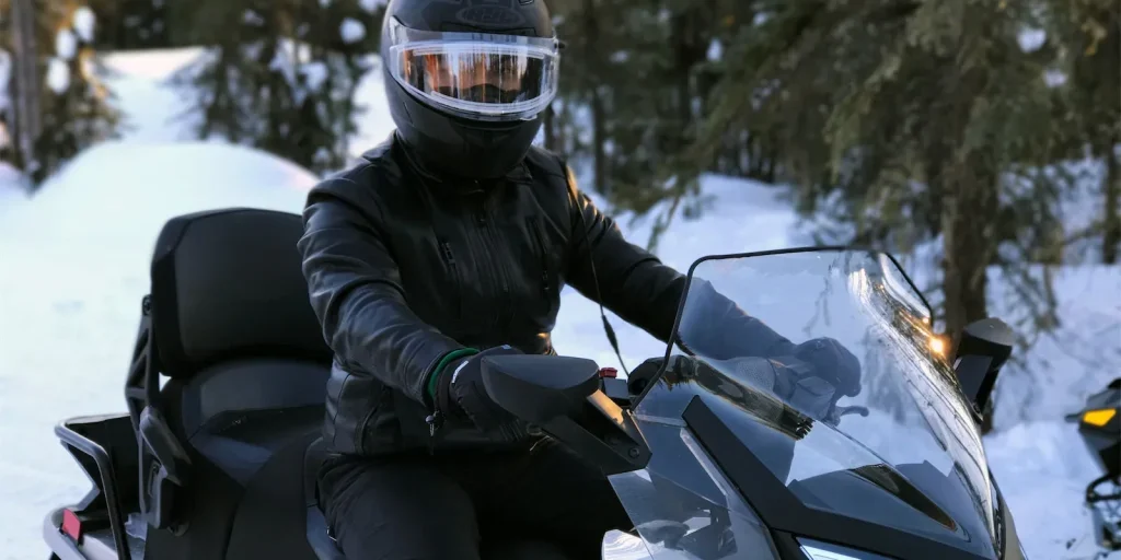 Person wearing a helmet and black leather jacket riding a snowmobile in a snowy forest setting, highlighting recreational vehicle use related to insurance coverage.